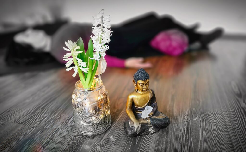Eine Buddha-Statue und weiße Blumen in einem Gefäß, im Hintergrund ist unscharf eine liegende Person zu sehen.