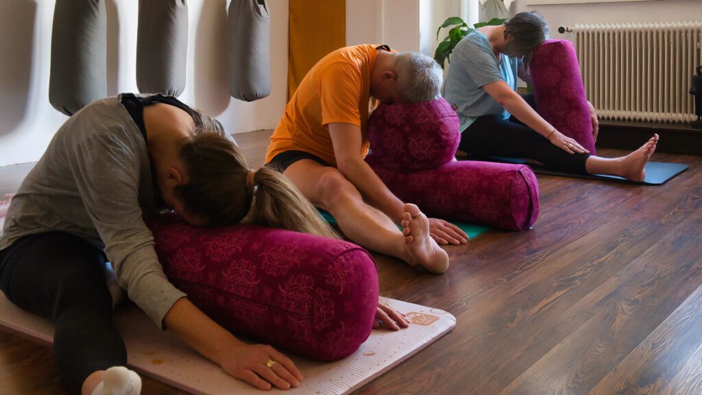 Drei Personen in einem Yogastudio machen sitzende Vorwärtsbeugen, jede von ihnen auf einem lila Nackenrollenkissen gestützt.