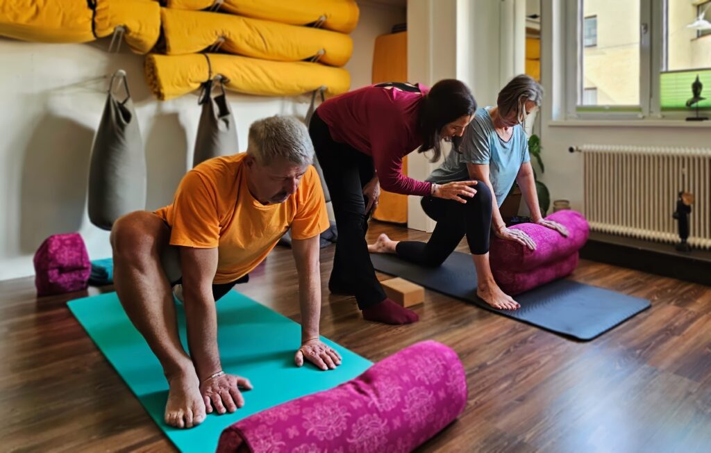 Drei Erwachsene praktizieren Yoga im Haus und verwenden dabei Polster, während ein Lehrer ihnen hilft und sie anleitet.