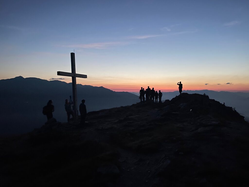 Silhouetten von Menschen in der Nähe eines großen Kreuzes bei Sonnenuntergang auf einem Berggipfel mit buntem Himmel im Hintergrund.