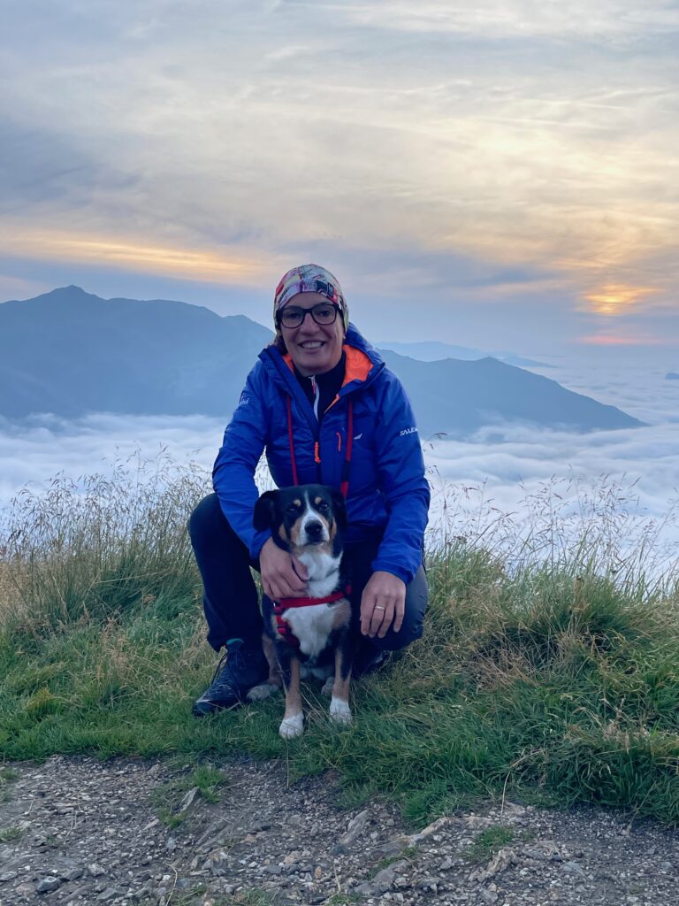 Person in blauer Jacke kniet bei Sonnenaufgang auf einem grasbewachsenen Hügel mit einem Hund, Bergen und Wolken im Hintergrund.