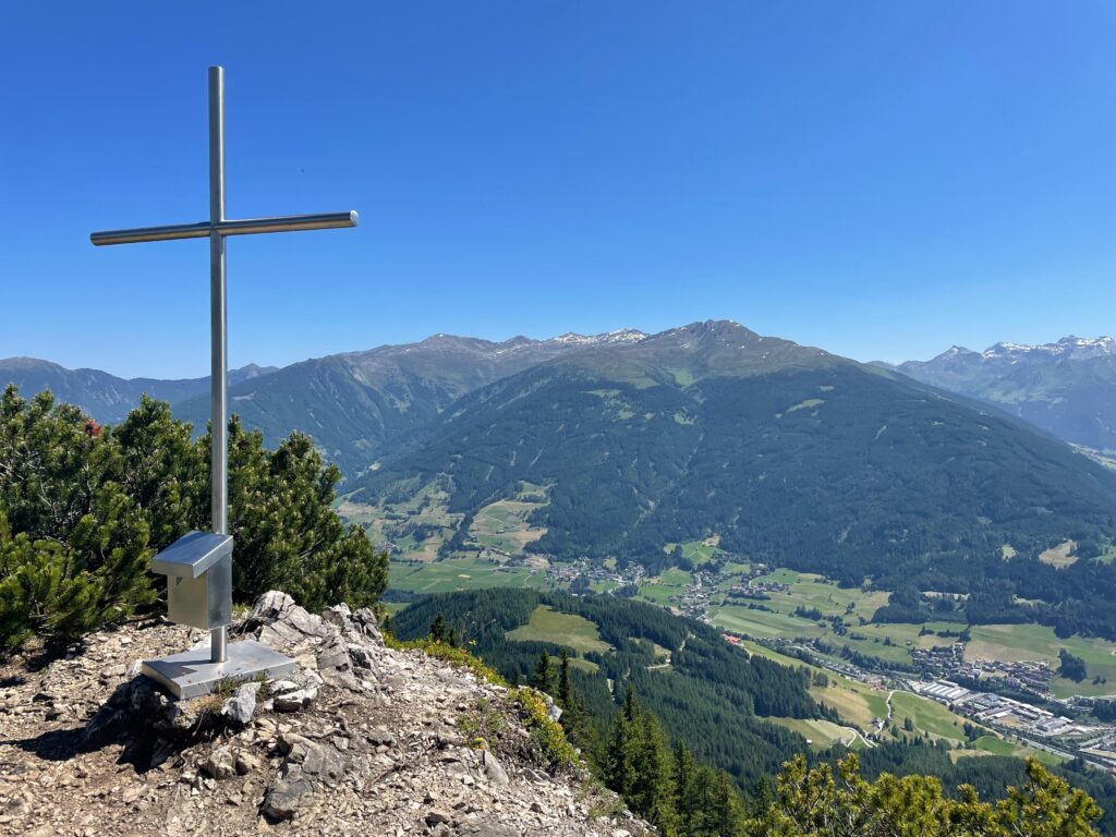 Auf einem Berggipfel steht ein Metallkreuz mit Blick auf ein Tal und ferne Berge unter einem klaren blauen Himmel.