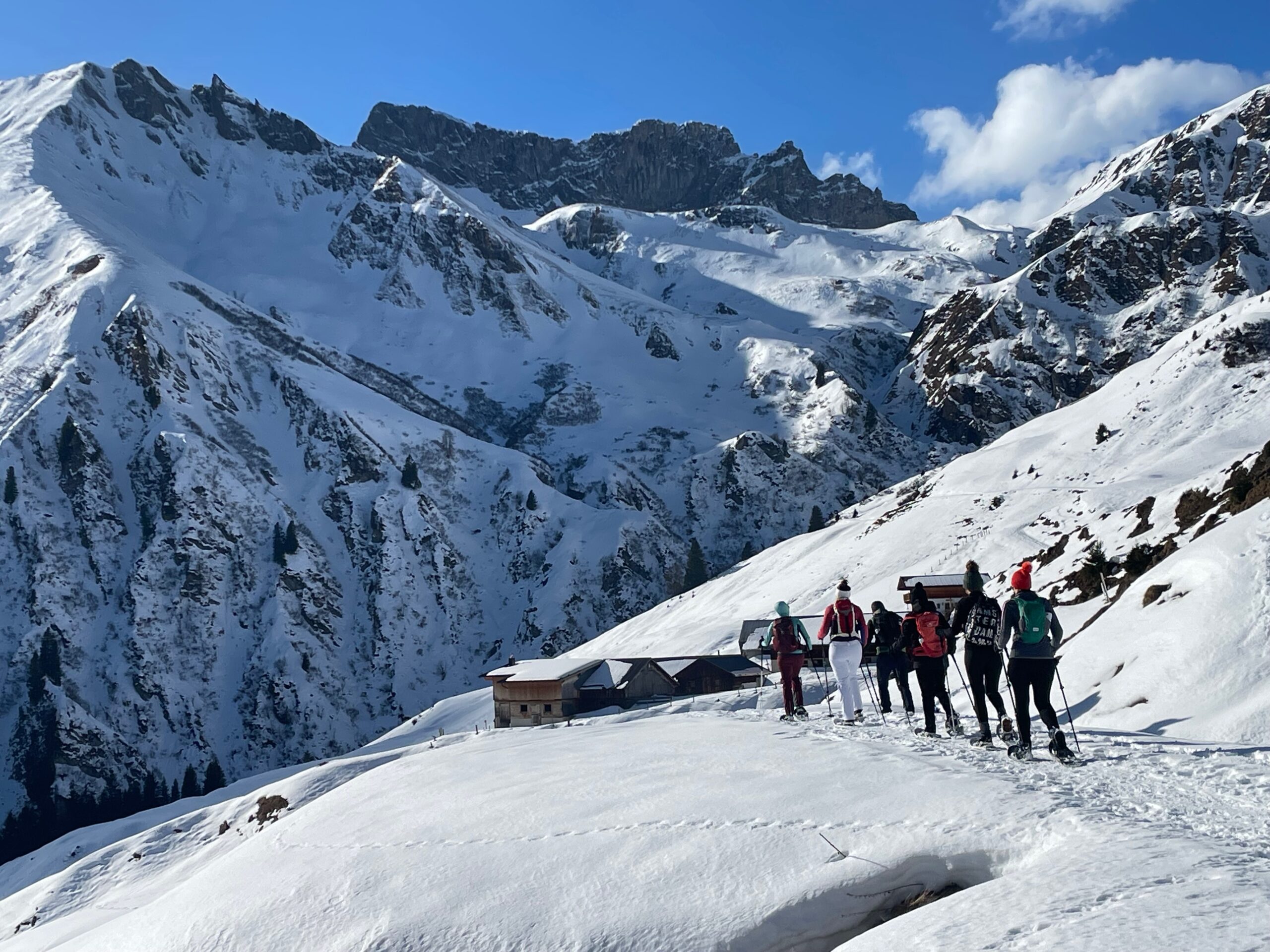 Sechs Personen wandern mit Schneeschuhen einen verschneiten Bergpfad hinauf, im Hintergrund sind hohe, schneebedeckte Gipfel zu sehen.
