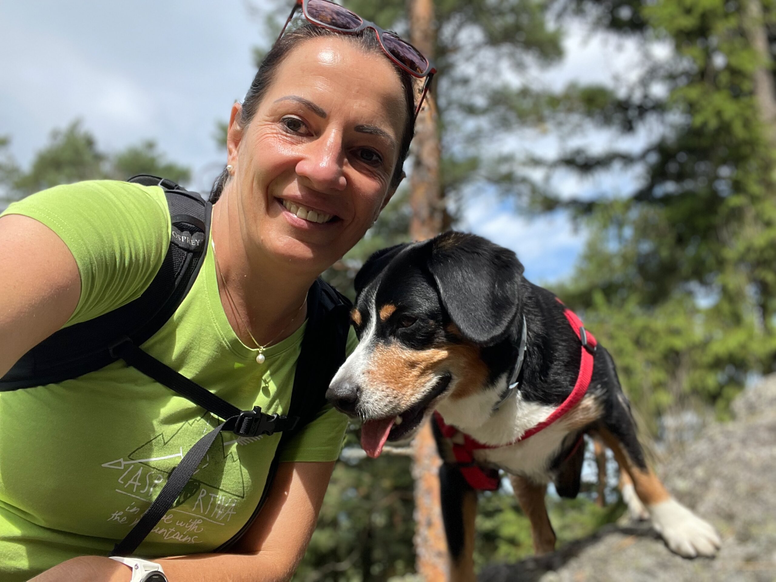 Frau in grünem Hemd lächelt im Freien mit einem schwarz-braun-weißen Hund in rotem Geschirr.