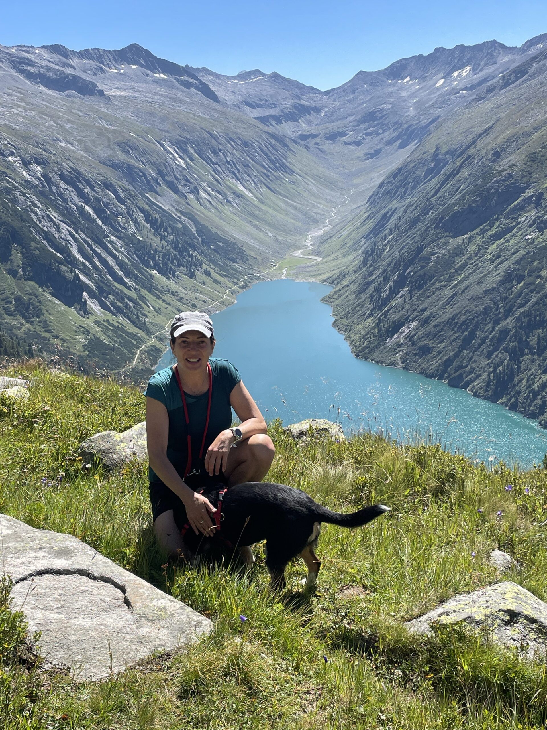 Bergwanderführerin Tirol Cornelia Rangger steht mit ihrem Hund auf einer grasbewachsenen Anhöhe mit Blick auf einen blauen Bergsee und schroffe Gipfel unter einem klaren Himmel.